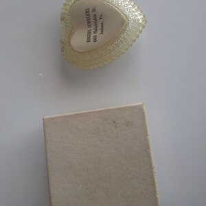 May include: A clear, heart-shaped trinket box with a textured, beaded edge and a cream-coloured interior. The box contains text that reads "ROGERS JEWELERS 660 Philadelphia St. Indiana, Pa." A small, beige cardboard box is also present.