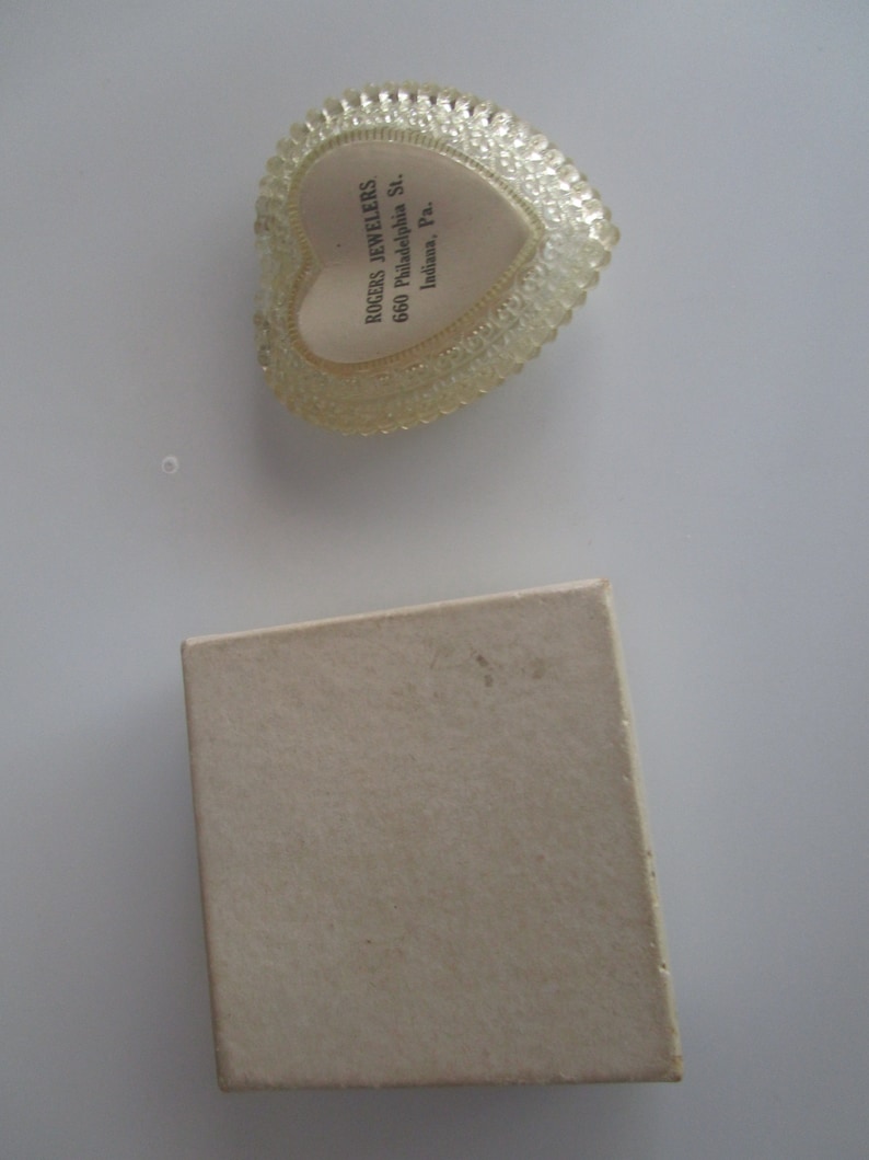 May include: A clear, heart-shaped trinket box with a textured, beaded edge and a cream-coloured interior. The box contains text that reads "ROGERS JEWELERS 660 Philadelphia St. Indiana, Pa." A small, beige cardboard box is also present.