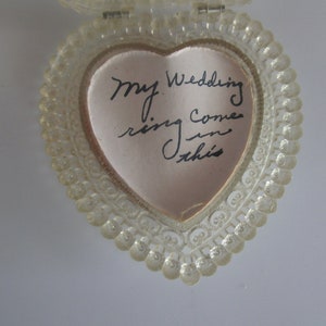May include: A clear, heart-shaped ring box with a beaded edge. Inside, a pink satin lining holds a handwritten note that reads, "My wedding ring comes in this."