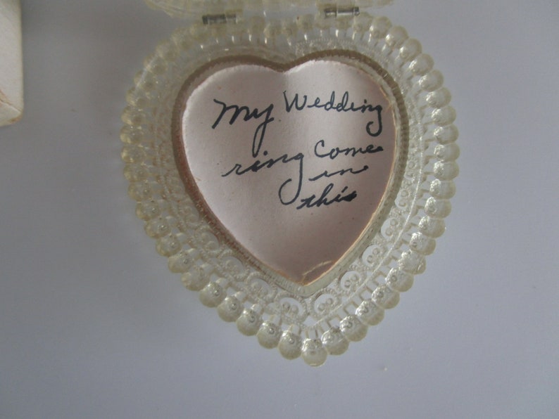 May include: A clear, heart-shaped ring box with a beaded edge. Inside, a pink satin lining holds a handwritten note that reads, "My wedding ring comes in this."