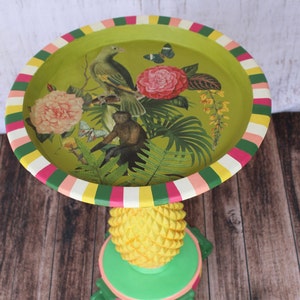 May include: A green and yellow pineapple-shaped side table with a green top featuring a tropical print of a parrot, monkey, and flowers. The top is surrounded by a multi-colored striped border.