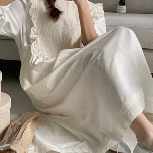 May include: A white cotton dress with a lace trim and a ruffled neckline. The dress has a loose fit and falls to the ankles. The model is wearing the dress with white shoes.