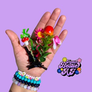 May include: A set of four black hair clips with a floral design. The design includes a red mushroom, a pink flower, a yellow flower, and a red apple. The hair clips are on a hand wearing several bracelets.