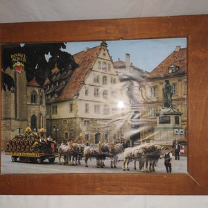 Peut inclure: Une photographie encadrée d'une charrette tirée par des chevaux transportant des fûts de bière devant un bâtiment historique avec l'inscription "Dinkel Acker" sur une pancarte. Le bâtiment est situé dans une ville européenne avec une statue au premier plan.