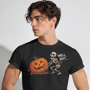 May include: A black t-shirt featuring a graphic of a skeleton pushing a large orange jack-o'-lantern pumpkin. The skeleton has a surprised expression and the text "WELL, SHIT...." is printed in white letters next to the skeleton's head.