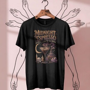 Può includere: Una t-shirt nera con una stampa grafica che riporta il testo "MIDNIGHT ESPRESSO BREWED UNDER THE FULL MOON". Il design include un bicchiere da martini, un teschio, candele, una pozione, cristalli e una luna crescente in tonalità oro e viola.