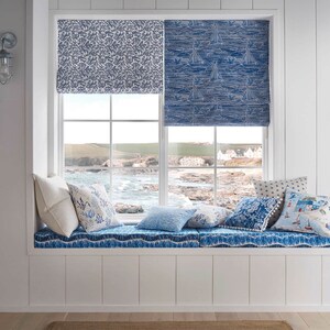 May include: A window seat with a white cushion and blue and white patterned pillows. The window has a view of the ocean and a rocky coastline. The window is covered with blue and white patterned roman shades.