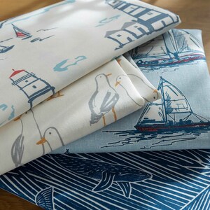 May include: Three fabric swatches with nautical themes. The top swatch features a white background with blue and gray illustrations of lighthouses, sailboats, and seagulls. The middle swatch has a light blue background with a dark blue sailboat and seagulls. The bottom swatch has a dark blue background with white stripes and a whale illustration.