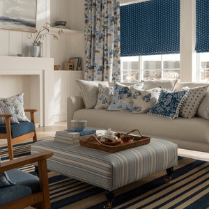 May include: A living room with a white sofa and blue and white patterned throw pillows. The sofa is in front of a window with blue and white patterned roman shades. There is a coffee table with a tray of teacups and a stack of books on it. The room has a blue and white striped rug.