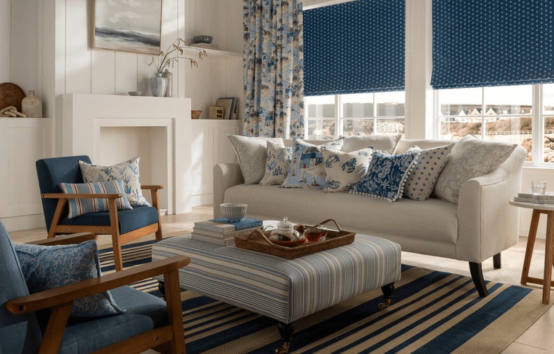 May include: A living room with a white sofa and blue and white patterned throw pillows. The sofa is in front of a window with blue and white patterned roman shades. There is a coffee table with a tray of teacups and a stack of books on it. The room has a blue and white striped rug.