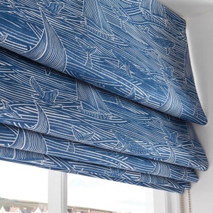 May include: Blue and white roman shade with a nautical pattern of whales, sailboats, and seagulls. The shade is made of a woven fabric and has a white cord.
