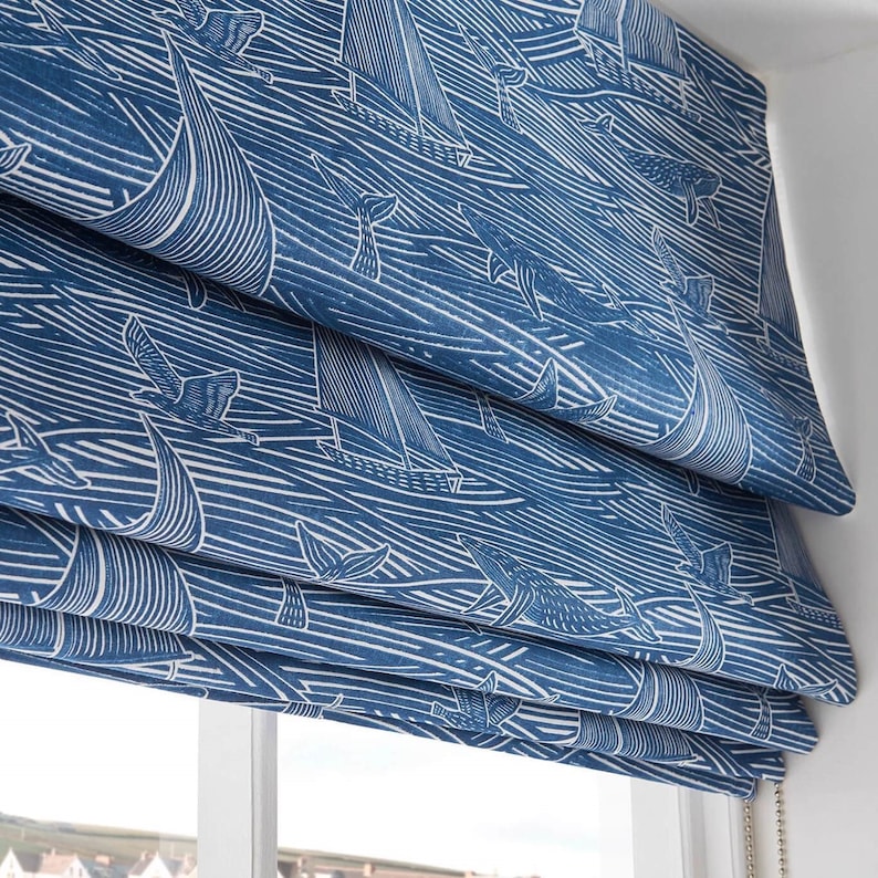 May include: Blue and white roman shade with a nautical pattern of whales, sailboats, and seagulls. The shade is made of a woven fabric and has a white cord.