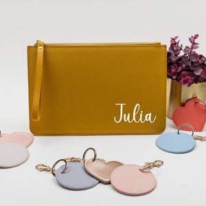 Op de afbeelding: Een mosterdgele leren clutch met een ritssluiting en een polsbandje. De clutch heeft de naam "Julia" in wit op de voorkant. De afbeelding toont ook verschillende sleutelhangers met verschillende gekleurde leren cirkels en harten.