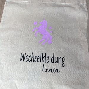 Op de afbeelding: Een beige tote bag met een roze eenhoorn en sterren ontwerp. De tas heeft zwarte tekst die "Wechselkleidung Lenia" zegt.