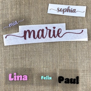 Op de afbeelding: Vijf strijklabels met verschillende namen. De namen zijn "mia", "marie", "sophia", "lima" en "paul". De labels zijn op een bruine jute achtergrond.
