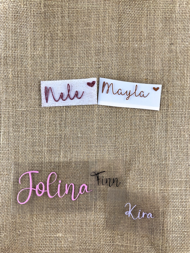 Op de afbeelding: Vier strijklabels met verschillende namen. De namen zijn "Nele", "Mayla", "Jolina" en "Kira". De labels zijn op een jute achtergrond.