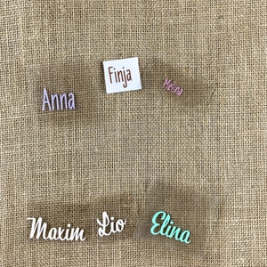 Op de afbeelding: Vijf transparante plastic naamstickers met verschillende namen erop geschreven in verschillende kleuren. De namen zijn Anna, Finja, Melina, Maxim, Lio en Elina. De naamstickers zijn op een bruine jute achtergrond.