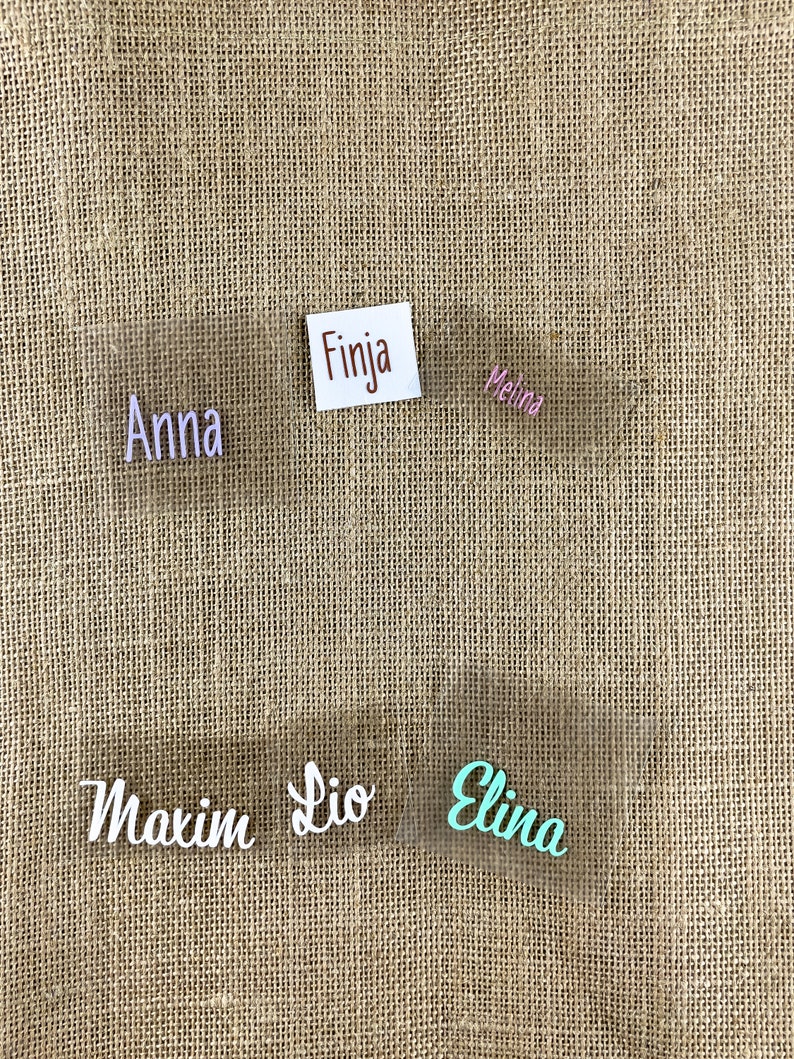 Op de afbeelding: Vijf transparante plastic naamstickers met verschillende namen erop geschreven in verschillende kleuren. De namen zijn Anna, Finja, Melina, Maxim, Lio en Elina. De naamstickers zijn op een bruine jute achtergrond.