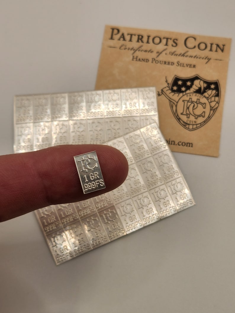 Op de afbeelding: Een zilveren Patriots Coin bar met de tekst "1 GR 999FS" erop. De bar wordt vastgehouden door een vinger.