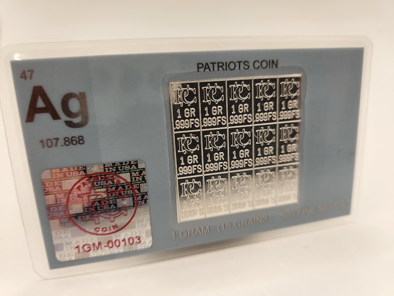 Puede incluir: Una barra de moneda de plata Patriots con una cuadr&iacute;cula de 16 cuadrados, cada uno con la inscripci&oacute;n "1 GR 999FS". La barra est&aacute; encerrada en un soporte de pl&aacute;stico transparente con un fondo gris. El soporte tambi&eacute;n presenta el s&iacute;mbolo qu&iacute;mico de la plata, "Ag", y el peso at&oacute;mico, "107,868". El texto "PATRIOTS COIN" est&aacute; impreso en la parte superior del soporte. El texto "1 GRAM (15 GRAINS) 999 FINE SILVER" est&aacute; impreso en la parte inferior del soporte.