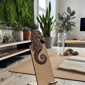 May include: A light-colored wooden phone stand featuring a dark, laser-engraved seahorse illustration on the front panel. The stand has a two-piece interlocking design with a small ledge at the bottom to support a mobile device in an upright position.