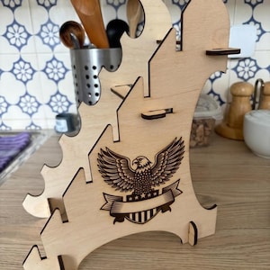 Könnte beinhalten: Ein hellfarbener Küchenutensilienhalter aus Holz mit einem Adleremblem. Der Halter hat mehrere Schlitze und ein dekoratives Ausschnittdesign. Er besteht aus hellfarbenem Holz und ist zur Aufnahme von Küchenutensilien bestimmt.