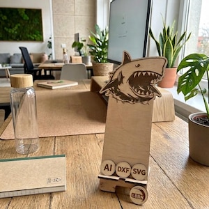 May include: A light-colored wooden phone stand featuring a laser-engraved shark head at the top. The stand includes three small circular wooden discs labeled AI, DXF, and SVG, and a circular logo disc at the bottom with the text LBRN.