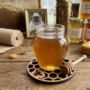 May include: A clear glass jar filled with golden honey sits on a circular wooden coaster featuring a laser-cut honeycomb pattern and a small bee icon. A wooden honey dipper rests beside the jar on the coaster, which is placed on a rustic wooden surface.