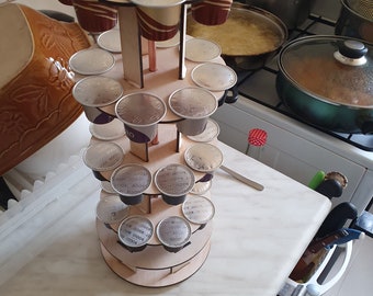 May include: A tiered wooden coffee pod holder with a circular design, featuring multiple levels to store individual coffee capsules. The structure is made of light-coloured wood and holds various coffee pods with silver foil lids in a vertical arrangement.