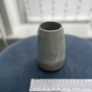 May include: A light grey ceramic vase with a speckled glaze. The vase has a rounded bottom and a slightly flared opening.
