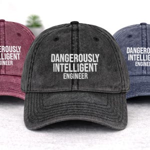 May include: Three baseball caps in shades of burgundy, charcoal gray, and navy blue. Each cap features the white text "DANGEROUSLY INTELLIGENT ENGINEER" across the front. The caps have a classic, worn look.