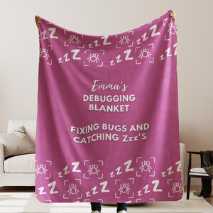 May include: Pink fleece blanket with a white bug and "zzz" pattern. The text "Emma's Debugging Blanket Fixing Bugs and Catching Zzz's" is printed in white on the blanket.