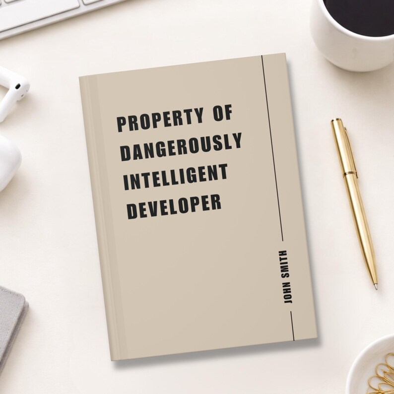 May include: A beige notebook with the text "PROPERTY OF DANGEROUSLY INTELLIGENT DEVELOPER" in black. The name "JOHN SMITH" is printed vertically on the right side. A gold pen and a cup of coffee are also visible.