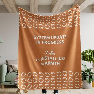 May include: A brown fleece blanket with a white repeating pattern of arrows. The blanket has the text "SYSTEM UPDATE IN PROGRESS" and "John IS INSTALLING WARMTH" printed on it.