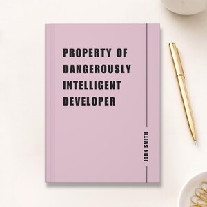 May include: A pale pink notebook with the text "PROPERTY OF DANGEROUSLY INTELLIGENT DEVELOPER" in black. The name "JOHN SMITH" is printed vertically on the right side. A gold pen and a white ceramic cup are in the background.