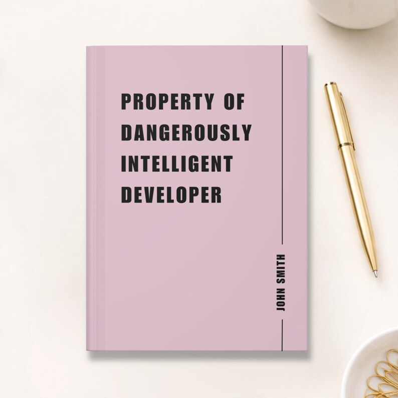 May include: A pale pink notebook with the text "PROPERTY OF DANGEROUSLY INTELLIGENT DEVELOPER" in black. The name "JOHN SMITH" is printed vertically on the right side. A gold pen and a white ceramic cup are in the background.