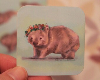 Puede incluir: Una pegatina con un wombat de dibujos animados que lleva una corona de flores. El wombat es marrón y tiene la nariz rosa. El fondo es azul claro con un tono verde.