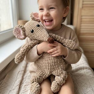 May include: A tan crocheted mouse plush toy with black eyes, pink inner ears, and a brown nose. The stuffed animal is held by a person wearing a tan sweater. The toy has long arms and legs and is made of a soft, textured yarn.