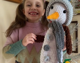 May include: A crocheted snowman doll with a gray hat, scarf, and brown arms. The snowman has a carrot nose and black eyes. The doll is white with gray and blue accents. The girl is wearing a pink sweater and blue jeans.
