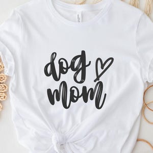 Può includere: Maglietta bianca con testo nero che dice "dog mom" con un simbolo di cuore.