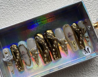 May include: A set of press-on nails in a silver box. The nails feature a variety of designs, including gold metallic accents, cherub and cross illustrations, and pearl embellishments. The nails are in a coffin shape and are labeled with an "M".