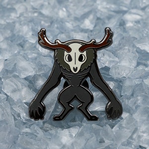 May include: A black and brown enamel pin of a creature with antlers, a skull-like face, and a furry body. The pin is on a background of blue ice crystals.