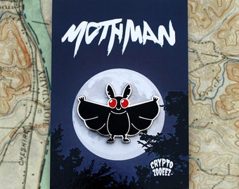May include: A black enamel pin featuring a mothman with red eyes, flying in front of a full moon. The pin has the text "MOTHMAN" at the top and "CRYPTO 200FEZ" at the bottom.