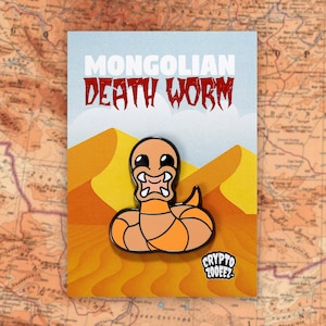 May include: A cartoon Mongolian Death Worm enamel pin with a black outline and orange body. The pin is set against a background of orange sand dunes. The text "MONGOLIAN DEATH WORM" is printed in white and red letters at the top of the card. The text "CRYPTO ZOOEEZ" is printed in white letters on a black background at the bottom of the card.
