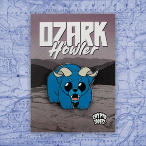 May include: A blue enamel pin featuring a cartoon monster with horns and large eyes. The text "OZARK Howler" is above the monster and "CRYPTO 200FEZ" is below.