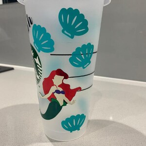 May include: A white reusable cup featuring a mermaid design. The mermaid has red hair and is wearing a purple seashell bra. The cup is decorated with teal seashells.