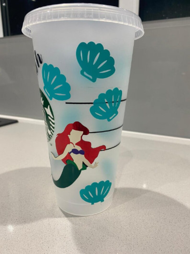 May include: A white reusable cup featuring a mermaid design. The mermaid has red hair and is wearing a purple seashell bra. The cup is decorated with teal seashells.