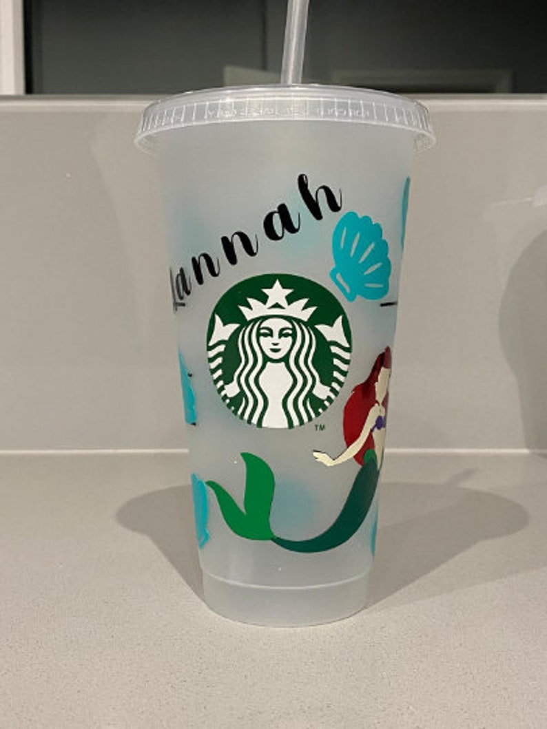 May include: A clear plastic cup with a mermaid design, a green Starbucks logo, and the name "Hannah" written on the side. The cup has a blue seashell and a green tail fin.