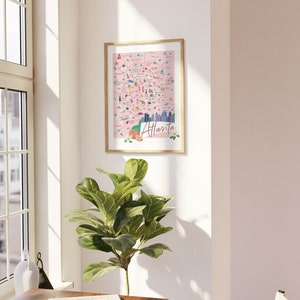 May include: A framed illustration of a pink map of Atlanta, Georgia with various landmarks and icons. The map is surrounded by a wooden frame and is hanging on a white wall.