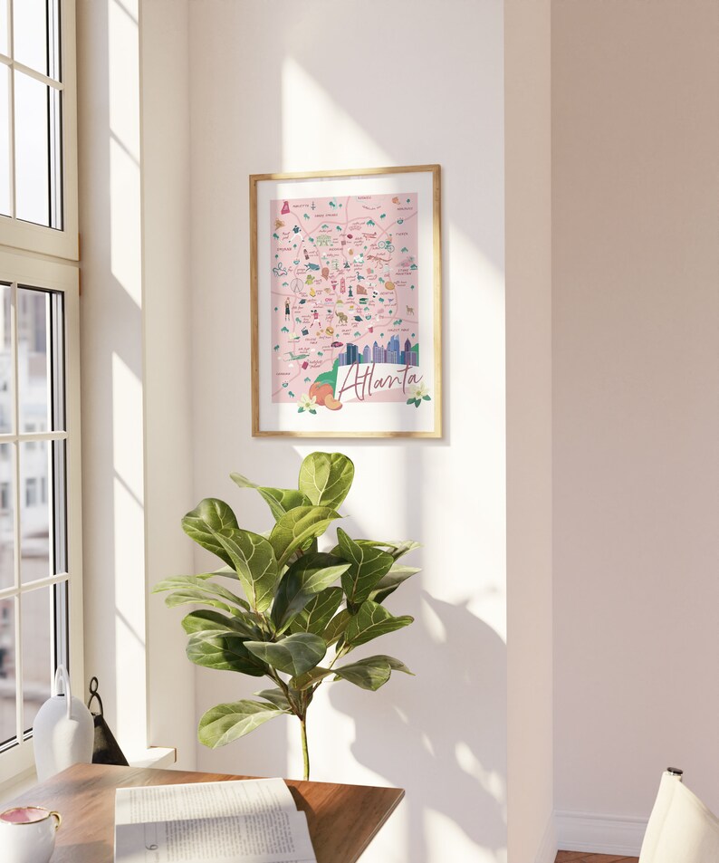 May include: A framed illustration of a pink map of Atlanta, Georgia with various landmarks and icons. The map is surrounded by a wooden frame and is hanging on a white wall.